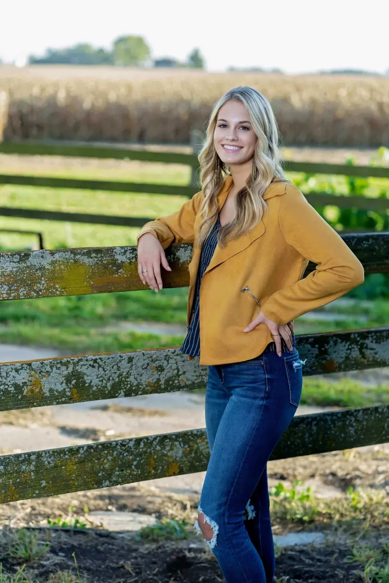 Teen girl in mustard yellow jacket leaning on weathered farm fence with hay bales and field in background, senior portrait