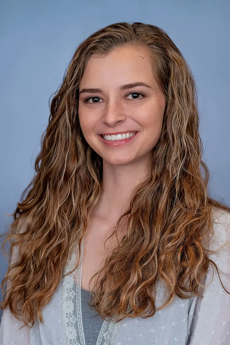 Professional studio headshot of woman with long curly hair in white lace top on light blue background