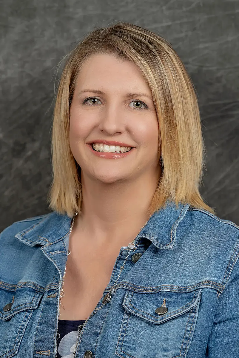 Professional studio headshot of smiling woman in denim jacket on gray textured background